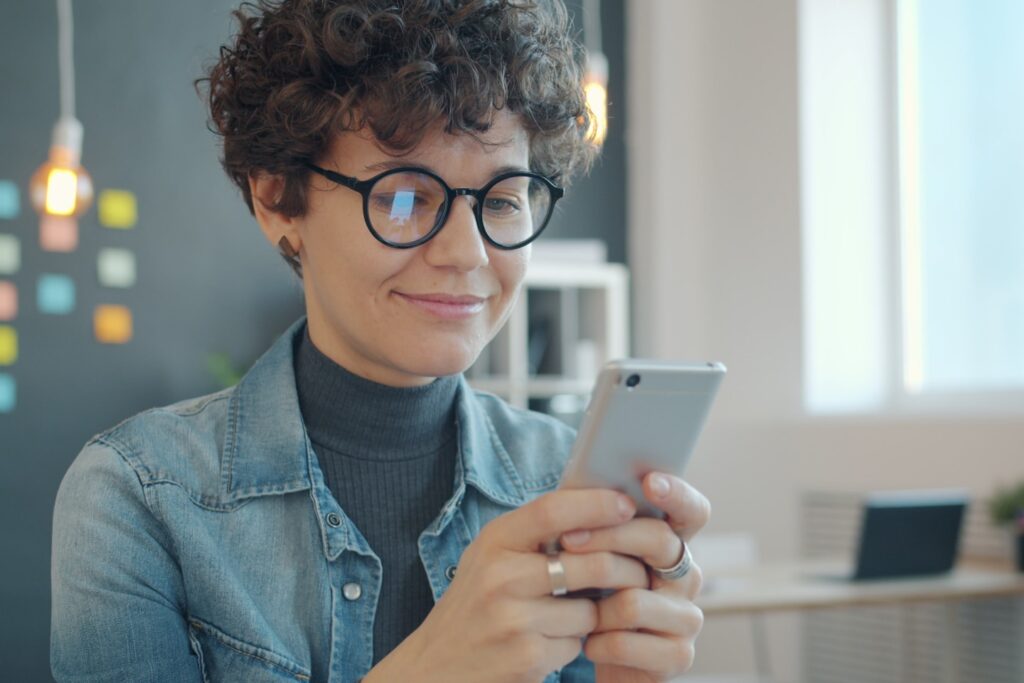 Smiling girl is using smartphone touching screen enjoying social media communication in workplace. Happy youth and modern technology concept.